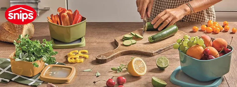 Contenedores SNIPS de colores con perejil, pimientos y fruta sobre una mesa de madera