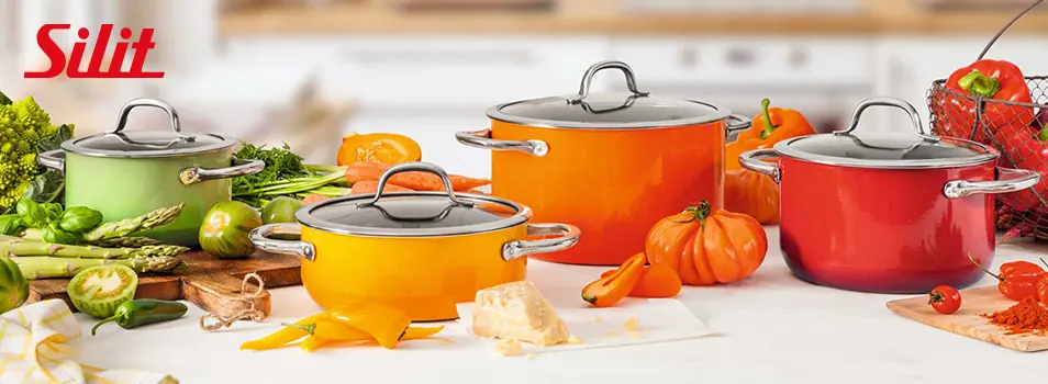 Ollas SILIT en colores verde, amarillo, naranja y rojo con tapas de vidrio rodeadas de verduras frescas