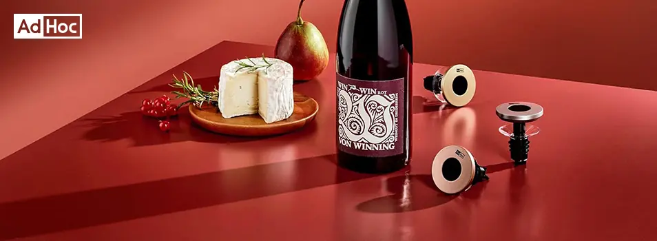 Tapones para botella AdHoc junto a una botella de vino tinto y un queso brie